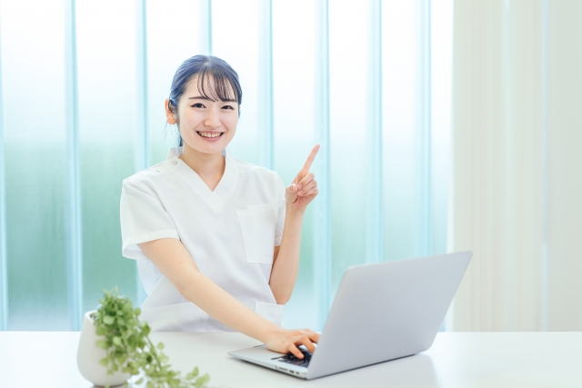 机の前に座っている白衣の看護師が人差し指を立てている。目の前にはパソコンが置かれている。