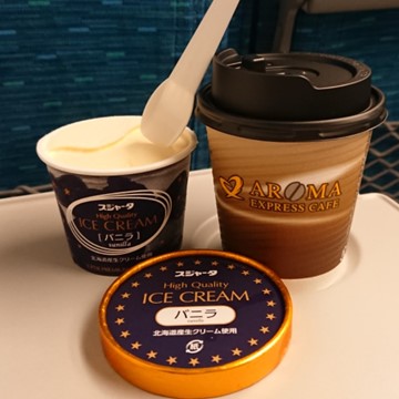 新幹線の車内のテーブルの上に、ふたのついた紙コップに入ったコーヒーと、新幹線の硬いアイスにスプーンがささっている。
